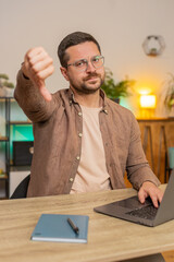 Dislike. Upset unhappy man using laptop and showing thumbs down sign gesture, expressing...