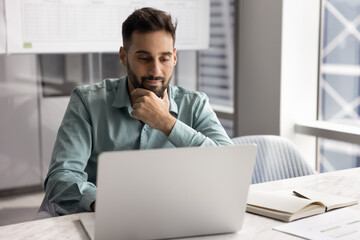Focused Latin businessman, office employee sits at desk look at laptop screen, watching on-line...