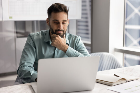 Focused Latin businessman, office employee sits at desk look at laptop screen, watching on-line training or professional webinar, reviewing business presentation, researching information for project