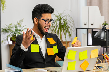 Tired exhausted Indian businessman working on laptop at office with many sticker task inscriptions. Freelancer man busy occupation feels sad boredom overworked multitasking. Panic attack, deadline