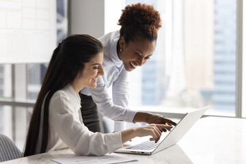 Two smiling multiracial women colleagues pointing at laptop screen together, laughing while reviewing amusing content, enjoy teamwork successful result during informal meeting or brainstorming session