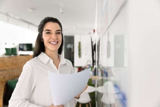 Latina professional woman, project coordinator or female office manager standing in creative coworking space, holds documents, reviewing wall calendar, posing for camera with contented smile, portrait