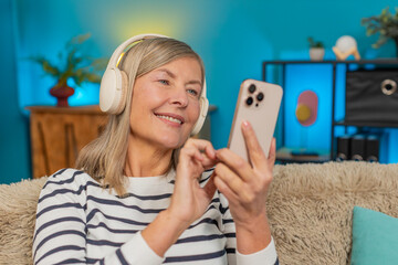 Mature woman creates music playlist on smartphone using wireless headphones while sitting on sofa...