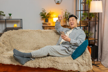 Happy overjoyed middle-aged man in wireless headphones relaxing lying on couch using phone at home choosing, listening favorite energetic disco dancing music. People weekend leisure activities.