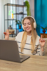 Caucasian woman at home on table wearing wireless headphones, typing on laptop, listening music,...