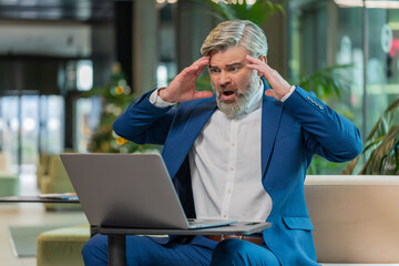 Sad middle-aged Caucasian businessman using laptop typing browsing, loses becoming surprised sudden...