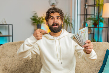 Successful rich young Indian man holding and waving dollar bill money fan sitting on sofa couch at...