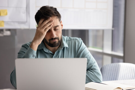Sad Hispanic man sit at desk distracted from laptop workflow with eyes closed, and hand against his forehead, experiencing head pain, feel stressed due to work pressure, deadlines, challenging tasks