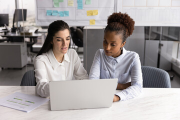 Two multiracial women colleagues sit at desk in office using laptop, discussing new task or...