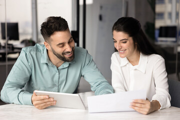 Two happy Hispanic colleagues reviewing data or reports displayed on digital tablet and paper documents, analyzing business performance metrics feel satisfied with progress, sales growth and success