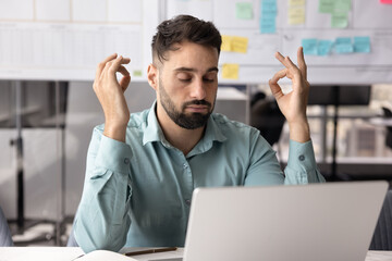 Male office employee sits at desk with laptop, take break, meditating with eyes closed, looks...