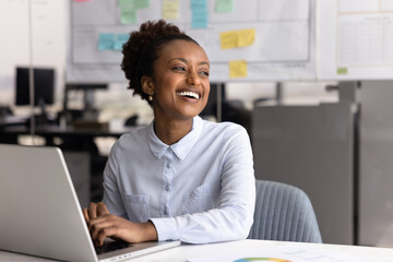 African woman sits at desk with laptop, looking away, engaged in creative tasks completion, feel...