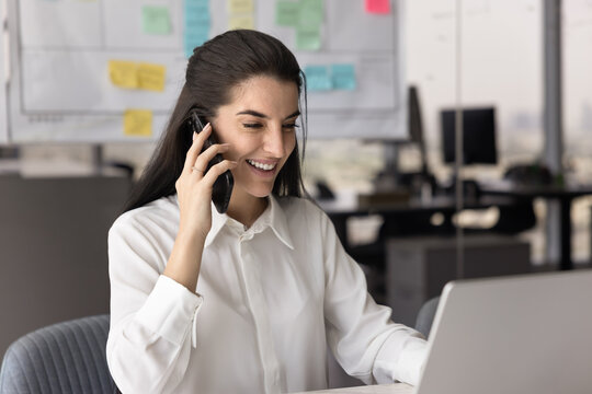 Smiling Latin businesswoman sit at desk in workplace, having positive conversation on smartphone while using laptop, provide information, consulting customer, discussing project with teammate remotely