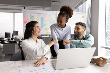 Three happy multiethnic workmates gathered in office working together on joint task, fist bumping...