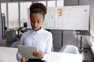 Serious African businesswoman using tablet standing alone in conference room, respond on client...