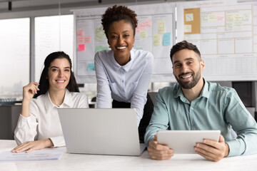 Portrait of three diverse colleagues posing for camera in office using laptop and tablet engaged in...
