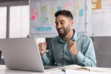 Happy Latin man sits at desk with laptop, celebrate completion of presentation or task...