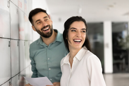 Two young, joyful Latin male and female colleagues laughing, looking away, engaged in task completion standing in modern coworking office. Positive workflow, teamwork and camaraderie among coworkers