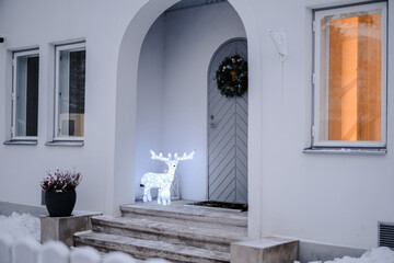 A charming house entrance adorned with festive winter decorations, featuring an illuminated glowing reindeer standing on the snow-covered steps
