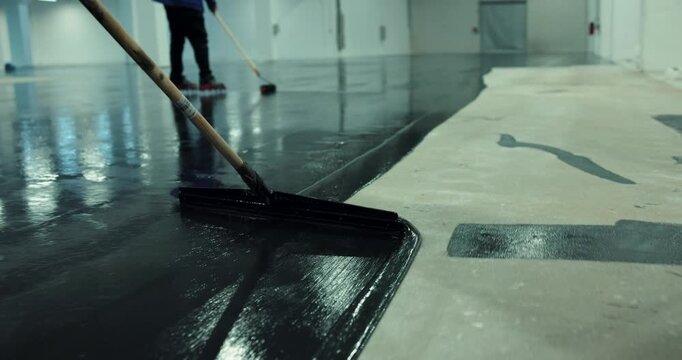 Workers using large squeegee and rollers to apply a dark epoxy coating to a concrete floor indoors