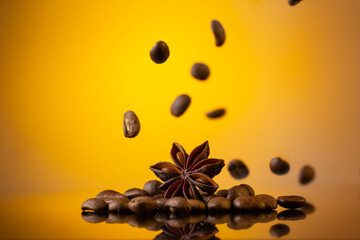 Coffee beans falling around star anise on a reflective surface with a yellow gradient background