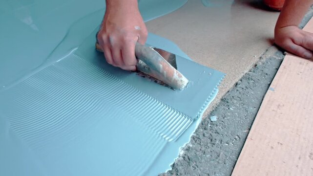 Closeup shot of a worker's hand uses a notched trowel to spread light blue epoxy primer on a floor