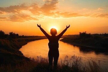 Fototapeta premium Silhouette of a person with raised arms standing by a riverbank during a vibrant orange sunset with scattered clouds and natural vegetation