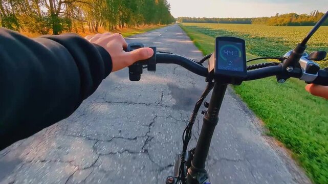 A man rides an electric bicycle on an asphalt road in summer. To the right of the road is a field. To the left of the road are birch trees growing in a row. The sun's rays are shining. POV
