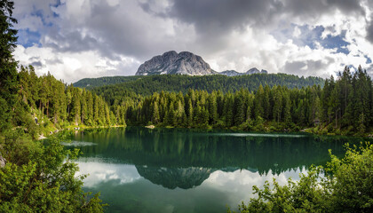 Panoramic view of mountain lake surrounded by dense forest under cloudy sky with moody light reflecting on calm water surface creating peaceful atmosphere