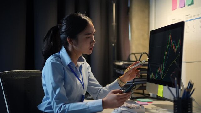 Young businesswoman analyzing financial charts displayed on a computer screen and smartphone, pointing at the monitor with a pen while sitting at her desk in a dimly lit office - Powered by Adobe