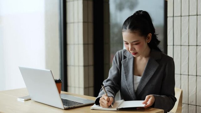 Confident professional businesswoman sitting at modern office desk, taking detailed notes in notebook while working intently on laptop computer during productive workday