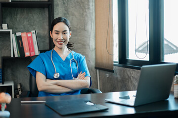 Smiling healthcare worker in blue scrubs with stethoscope, seated at desk in a contemporary office...
