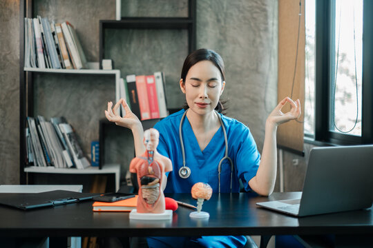 Female doctor in blue scrubs meditating at desk in a modern office, promoting mindfulness and stress relief in healthcare.