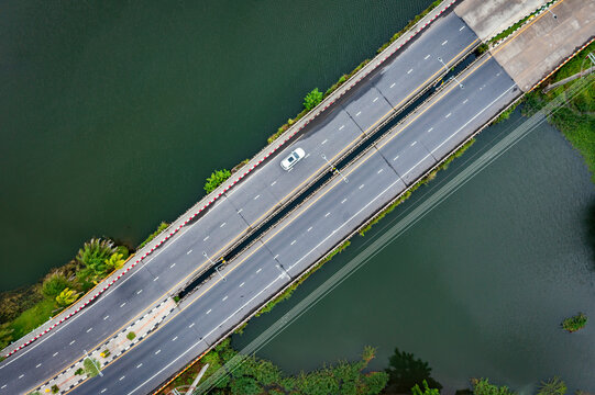 Aerial view of a long, modern highway crossing calm green waters, with a lone car traversing the straight path amidst lush nature.