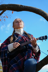 Beautiful blonde girl playing guitar ukulele and singing relaxing enjoying outdoors on sunny autumn...