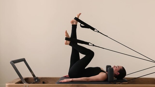 Slim brunette woman in black tight sportswear working out on Pilates reformer machine. Stretching and toning muscles during Pilates session in bright fitness studio.
