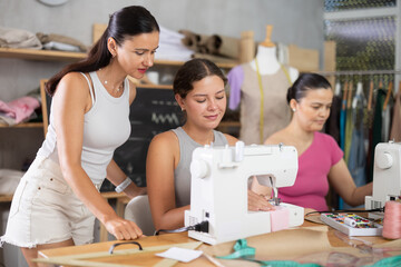 Enthusiastic girl in twenties stitching at machine in tailoring studio, enjoying hands-on learning during group sewing class for adults, while fellow learners sharing creative work and friendly