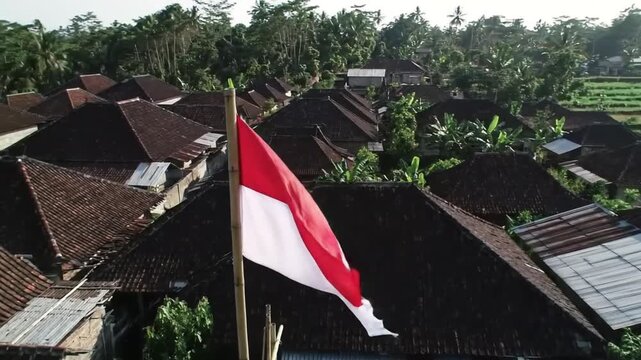 Indonesian flag on a pole flying above residential rooftops in a green village area