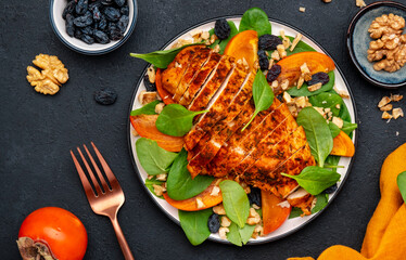 Autumn gourmet salad with fresh persimmon, grilled chicken fillet, walnuts, raisin and spinach, black background, top view