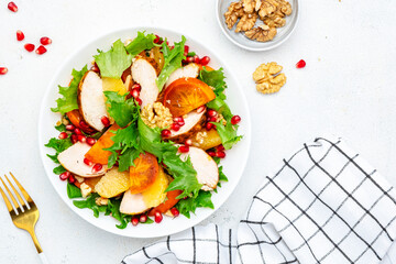 Autumn persimmon salad with grilled chicken fillet, pomegranate seeds, walnuts, oranges and lettuce, white table background, top view