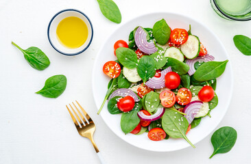 Healthy vegan vegetable salad with spinach, tomatoes, cucumber, onion, sesame seeds and olive oil. White table background, top view