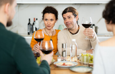 Friends are sitting at table, chatting sweetly, discussing and telling news, sharing plans. Married couple celebrates housewarming party with relatives.