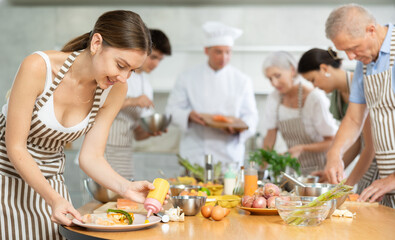 Pleased young female member of cooking club engaged in decoration of baked piece of salmon in the kitchen