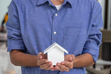 Close-up of man in blue shirt holding small white house model with both hands, symbolizing home...