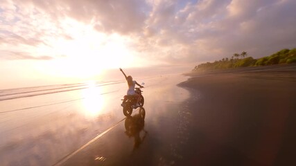 FPV drone shot of woman riding motorcycle along tropical beach during golden sunset, creating cinematic, free-spirited atmosphere. Concepts of freedom, adventure, and slow travel lifestyle.