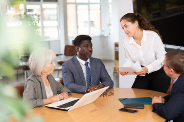 In conference room, young woman head of department speaks at meeting and reports unpleasant news to frowning colleagues. Secretary records leading moments of conversation in laptop