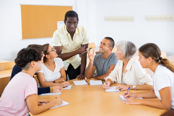 Positive friendly African American English tutor giving language courses to group of interested adults of different ages and nationalities sitting around table in classroom..