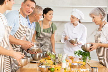 Smiling interested adult man attending group culinary class, standing by table with ingredients and utensils, learning culinary skills from professional chef
