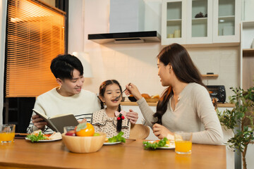 Smiling Asian family enjoying breakfast in weekend morning. parents and daughter, cooking, and sharing joyful at the dining table in the kitchen together.
