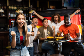 Cheerful excited young female fan watching sports game in bar, gesturing emotionally, happy with victory of favorite team against background of people with national flag of Germany..